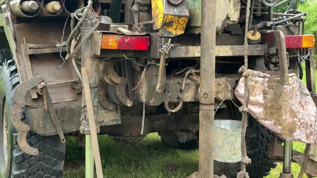 Tracked drilling rig extracting soil cores on grassy field, metal tracks visible, auger drilling into ground, columns of extracted soil stacked nearby, muddy site conditions under overcast.