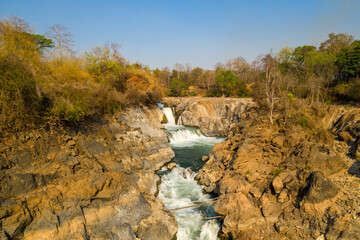 Warm sunlight illuminates the rugged rocky cascades and dry forest at Chutes de Khone on the Mekong River in southern Laos. The exposed stone and layered rapids create striking natural textures © Florent