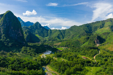 Fototapeta premium Sweeping view of a lush valley surrounded by dramatic limestone mountains and dense tropical forest near Nong Khiaw, Laos. Bright daylight highlights rice paddies, winding roads, and vibrant