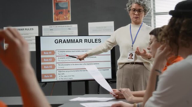 Medium shot of grey haired mature woman as female teacher pointing at digital screen and explaining English grammar to group of female inmates listening during class in prison education program