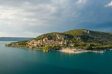 Fototapeta premium Sunlit village of Bauduen sits on a peninsula surrounded by turquoise waters, with a sandy beach and marina nestled beneath green hills and dramatic rocky outcrops under a cloudy sky.