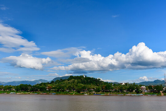 A panoramic view of the Mekong riverfront in Luang Prabang, Laos, with lush greenery, riverside houses, and Mount Phousi crowned by its golden stupa under a vibrant blue sky with dramatic cumulus