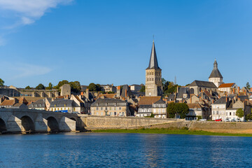 Fototapeta premium Warm evening sunlight highlights the medieval church spires, stone bridge, and traditional rooftops of La Charite sur Loire, with the calm Loire river in the foreground. The scene captures the