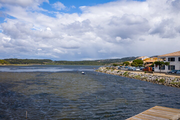 Obraz premium Row of waterfront houses and parked cars line a rocky embankment beside choppy lagoon waters in Gruissan. Dramatic clouds and sunlight create a dynamic sky over the tranquil village landscape.