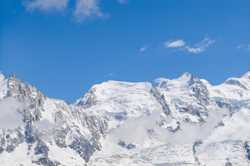Obraz premium Snow-capped peaks of Mont Maudit and Mont Blanc du Tacul rise above drifting clouds, set against a vibrant blue sky in the French Alps.