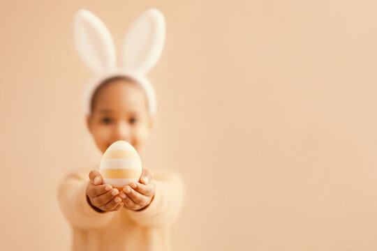 Black child wearing bunny ears holding striped Easter egg toward camera. Spring holiday celebration, childhood, pastel Easter concept with copy space