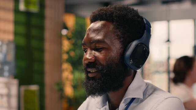 African american man in good mood listens to music on headset, working with laptop and combining stress relief with focused business analysis. Multitasking with audio and tasks in office.
