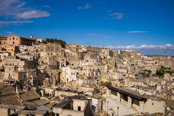 Expansive cityscape of Matera, Italy, showcases layered stone buildings and historic architecture bathed in sunlight beneath a vivid blue sky. The intricate textures and warm tones evoke a timeless