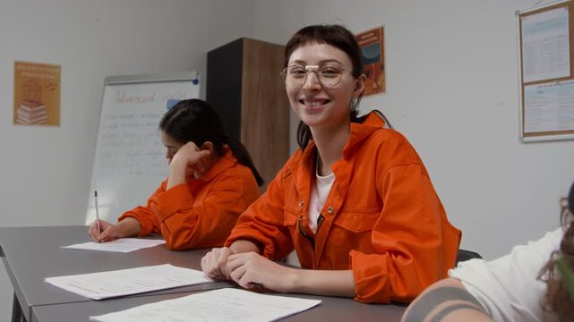 Zoom in shot of smiling young woman as female inmate looking at camera while sitting at desk with group of people doing tests during class in prison education program, copy space