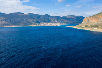 Naklejka premium Aerial view of vivid blue Mediterranean waters stretching toward a coastal village at the base of rugged mountains, with sunlit cliffs and open sky creating a striking landscape near Monemvasia.