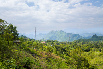 Vibrant hills covered in dense green foliage with a tall communication tower rising above the treetops. Layered mountains fade into the distance under a partly cloudy sky, creating a sense of depth