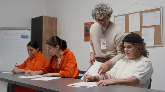Medium shot of group of female inmates sitting at desks in class doing tasks with grey haired female teacher assisting and checking work during lesson in prison education program, copy space