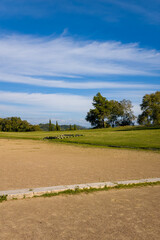 Expansive view of the ancient Olympic stadium in Olympia, Greece, featuring a sandy running track bordered by grassy embankments and scattered stone remains. Bright daylight and a vivid blue sky
