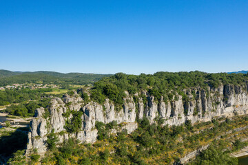 A rugged limestone ridge topped with dense green trees rises above the landscape in the Gorges de l'Ardeche, with clear sunlight highlighting the rock textures and a vivid blue sky overhead.