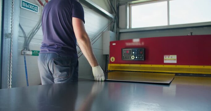 Industrial worker positioning metal sheet on guillotine shear before cutting operation. Factory environment highlighting blue-collar skills and manufacturing accuracy