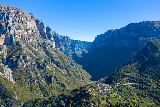 Wide aerial view captures the dramatic limestone cliffs of Vikos Gorge towering above a small mountain village in Epirus, Greece. Bright sunlight and clear blue sky highlight the rugged rock faces and