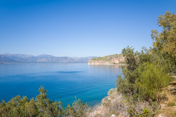 Obraz premium Tranquil coastal scene featuring crystal-clear blue water, lush green shrubs, and rugged cliffs under a bright sky. The serene Mediterranean landscape is framed by natural vegetation and distant