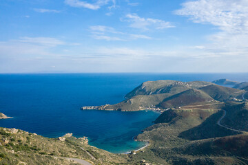 Fototapeta premium Aerial view of a sweeping bay surrounded by rugged hills and winding roads along the Magne coastline in Greece. Clear blue water contrasts with the arid, textured landscape under a bright sky.