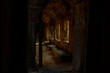 Obraz premium Dim natural light filters through stone windows, illuminating the textured walls and benches of a narrow corridor inside Bayon temple in Angkor, Cambodia. The weathered sandstone and carved details