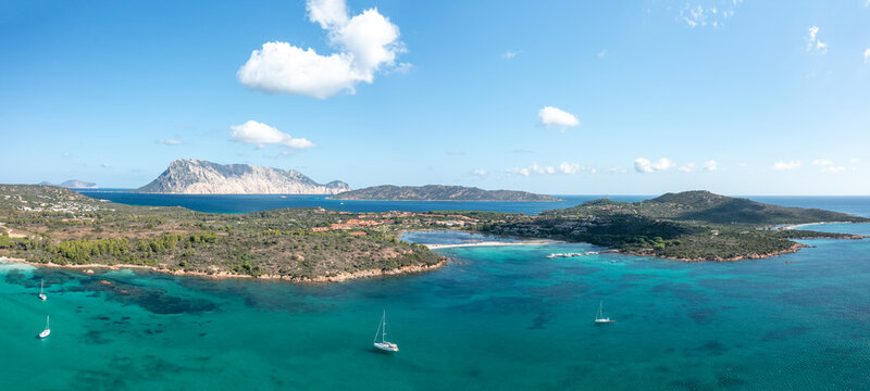 Aerial panorama of Spiaggia Salina Bamba near San Teodoro, Sardinia, featuring turquoise waters, anchored sailboats, rugged coastline, and the striking silhouette of Tavolara island under a bright