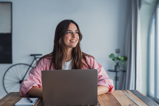 A young woman sits at her desk at home distracted from work, staring into the distance as she imagines new possibilities and enjoys a peaceful moment of reflection