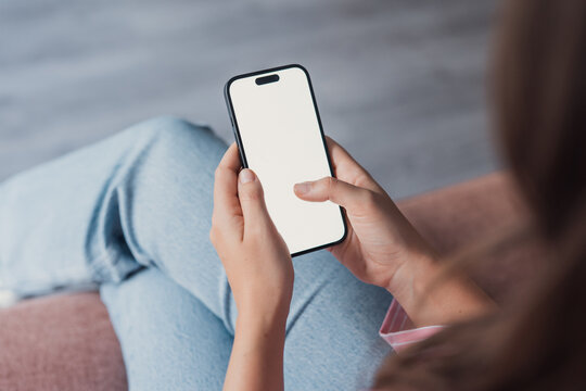 A millennial woman rests indoors while using her mobile phone with a mockup screen, reading updates, calling, or messaging, capturing everyday technology use in a comfortable home setting