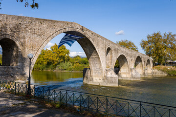 Fototapeta premium Historic multi-arched stone bridge stretches across a calm river in Arta, Greece, with a large Greek flag suspended beneath one of the central arches. Autumn trees and textured masonry are illuminated