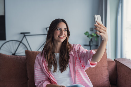 Happy pleasant millennial woman relaxing on comfortable couch, holding smartphone in hands. Smiling young lady taking a selfie, posting on social media, using mobile applications at home.