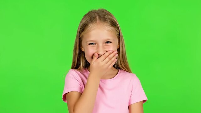A joyful young girl wearing a pink t-shirt is captured laughing and covering her mouth with her hand, isolated on a green screen background