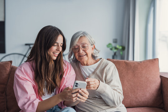 Smiling older mother and adult daughter using phone together, sitting on cozy sofa at home, happy young woman and mature mum looking at smartphone screen, watching video, having fun, two generations