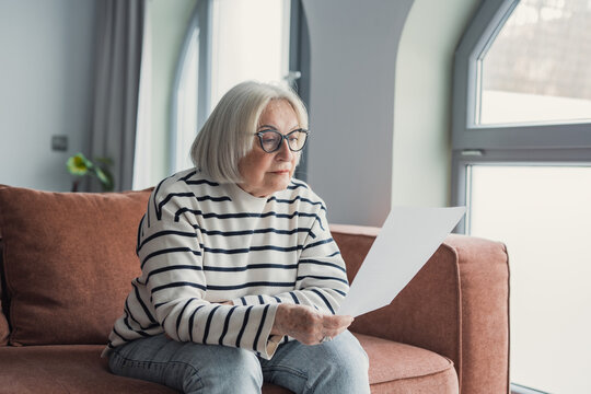 Focused elderly 60s woman sit on sofa in living room reading postal correspondence or bank notice, concentrated mature 50s female rest on couch at home consider message in paper post letter
