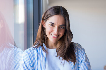 Obraz premium A positive young student or professional poses near a window with a natural smile, presenting a bright headshot that conveys optimism, growth, and readiness for new opportunities
