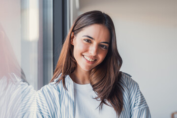 A happy businesswoman stands by a window smiling warmly at the camera, creating a professional portrait that reflects ambition, approachability, and career confidence © Daniel