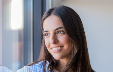 Fototapeta premium A young woman stands by the window in winter, enjoying warm sunlight and looking outside with a calm and relaxed expression, capturing a peaceful moment of comfort and quiet reflection at home