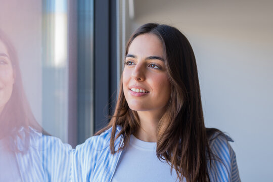 Standing near a window, a young woman gazes at the winter scenery as soft sunlight reflects on her face, showing happiness, serenity, and a gentle lifestyle moment indoors