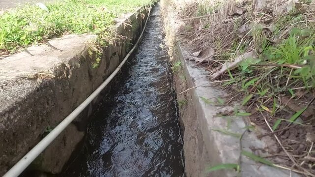 Clear water flowing through a concrete drainage ditch with a white irrigation pipe in a rural area.