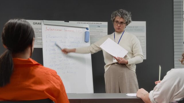Medium shot of grey haired female teacher leading life skills class and pointing at board with interview tips while group of inmates listening and taking notes during prison education program
