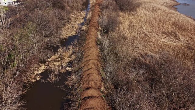 A long earthen berm is being built beside a waterway in a rural setting. The area shows trees and tall grass with water visible in the foreground.