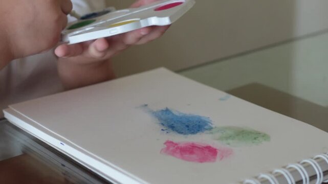Detalle de manos infantiles pintando con acuarelas de colores sobre un cuaderno de dibujo en casa. Concepto de creatividad, educaci&oacute;n art&iacute;stica e infancia.