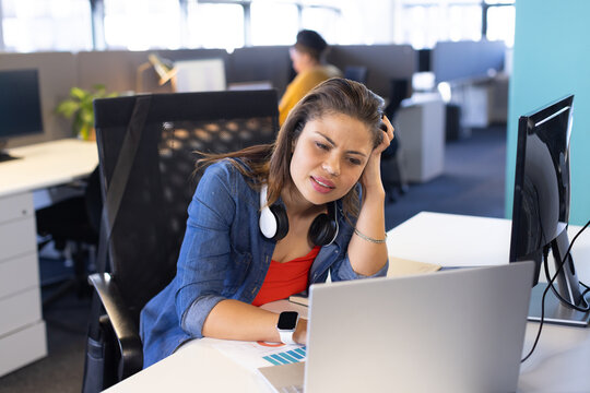 Diverse female coworkers sitting at office desk focusing on laptop with charts, headset, smartwatch