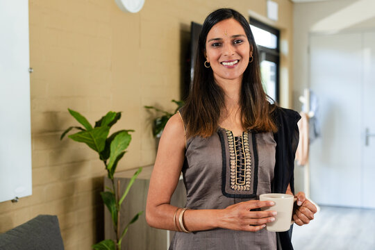 Indian adult woman standing in lounge holding white mug wearing kurti, black shawl with whiteboard