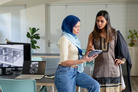 Indian female colleagues collaborating over floor plans using tablet and monitor in design studio