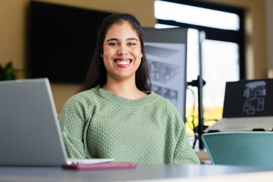 Smiling Hispanic woman sitting at light-colored table in design studio with laptop and floor plans