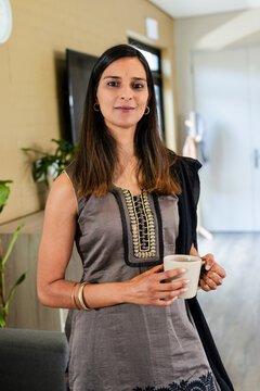 Indian woman wearing grey kurti holding ceramic mug while standing in modern home living area