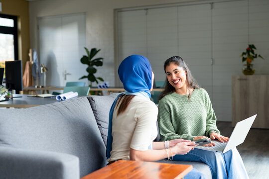Diverse women coworkers sitting on sofa in office using smartphone, laptop, monitor, rolled papers