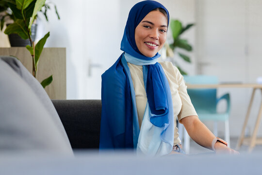 Indian Asian woman wearing blue headscarf sitting on grey sofa with potted plant in office lounge
