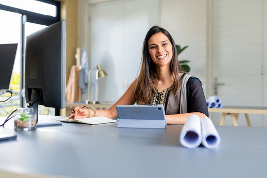 Indian woman sitting at office desk organizing architectural blueprints by computer and tablet