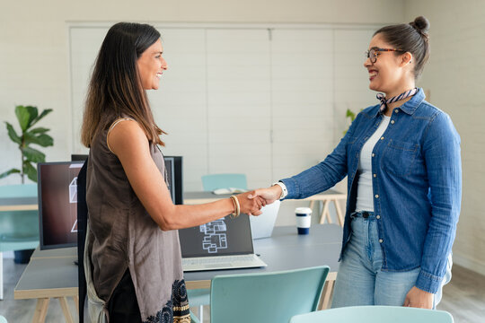 Diverse female coworkers shaking hands over floor plans on office table with laptop and coffee cup