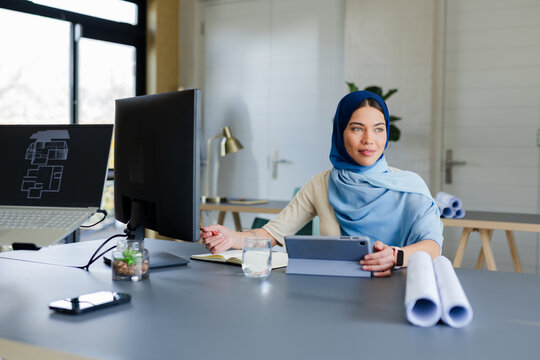 Indian Asian woman sitting at desk in workspace reviewing blueprints on dual monitors with laptop