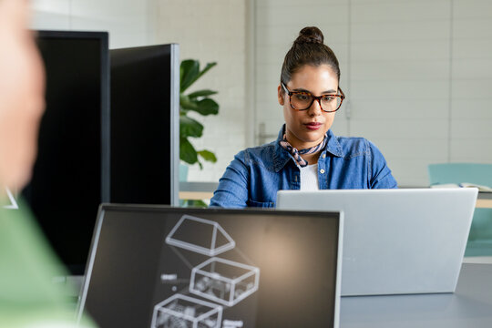 Female tech professional wearing glasses working at office desk using silver laptop and 3D monitor